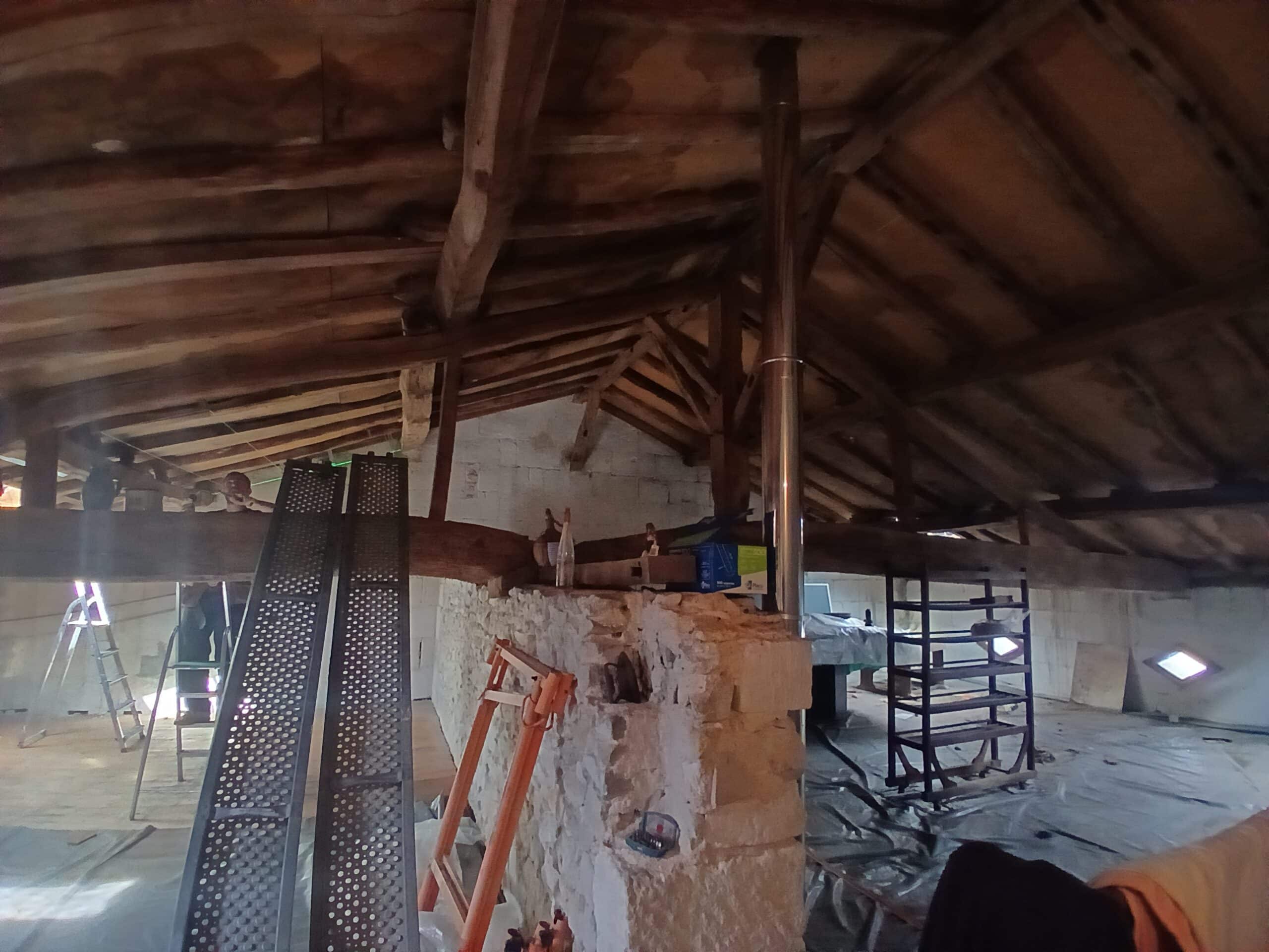 Rénovation d'un grenier avec charpente bois et conduit de cheminée en inox, équipements de chantier visibles.