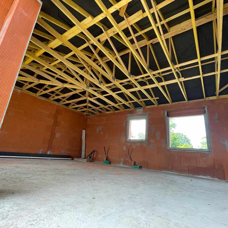 Intérieur d'un chantier de construction avec murs en briques rouges, plancher béton brut et charpente bois apparente sous une membrane noire.