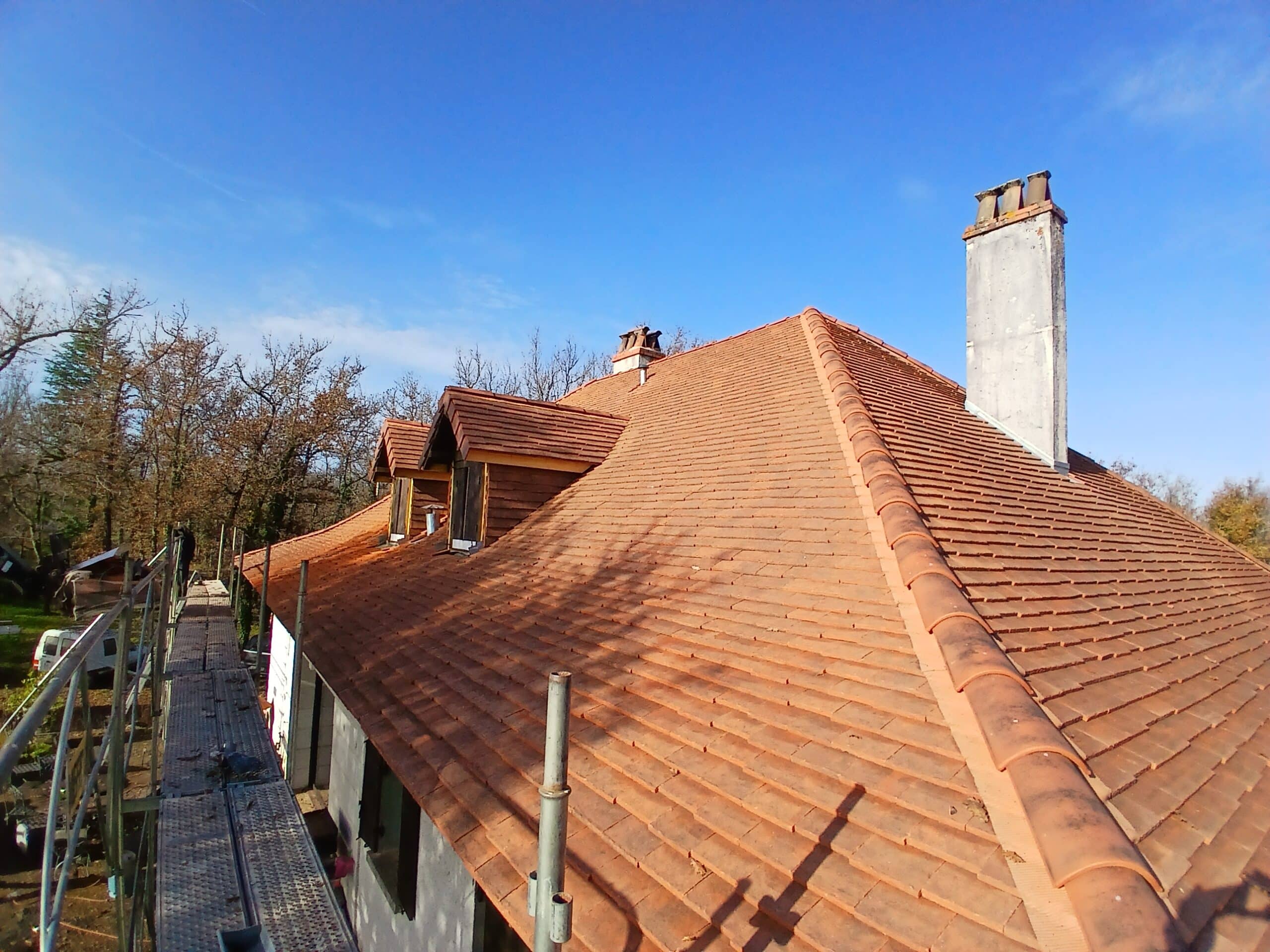 Toiture en tuiles rouges neuves, échafaudage visible, gros conduit de cheminée et lucarnes sous un ciel bleu dégagé.
