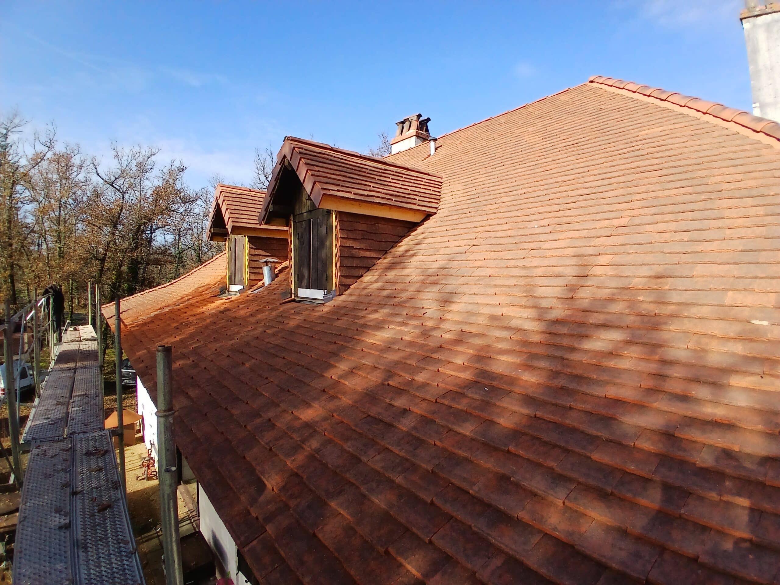 Toit en tuiles terre cuite neuves sur maison avec lucarnes en bois, échafaudage visible, ciel bleu.